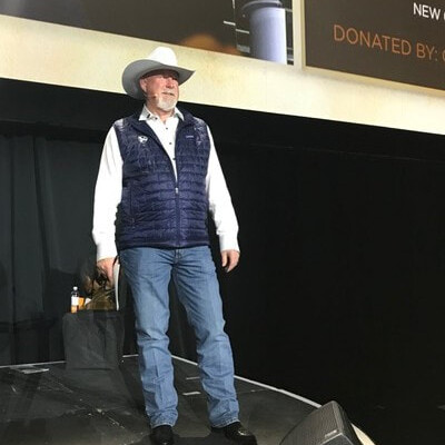 man stands on stage at a fundraising event wearing a cowboy hat