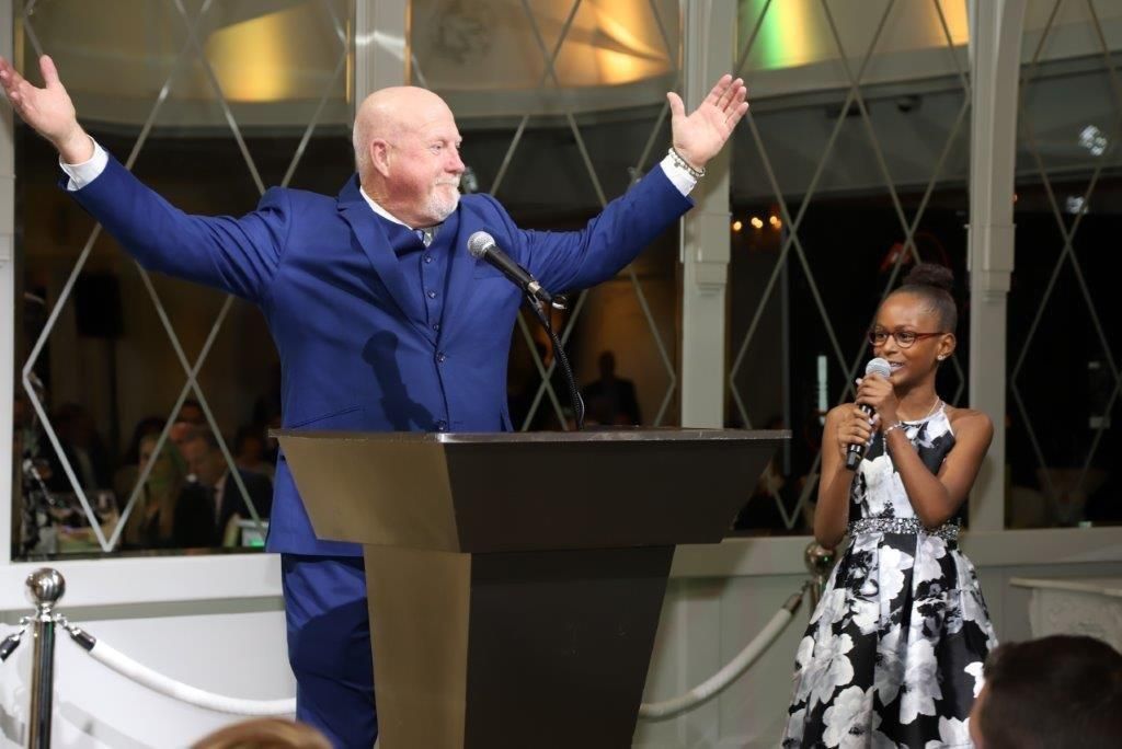 man in suit stands at podium with girl speaking into microphone during an auction